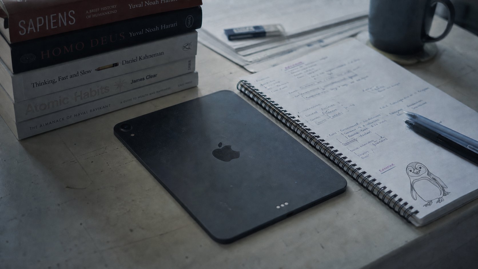 サピエンス全史などの本の山の隣に伏せて置かれた iPad、隣のノートにはペンギンの落書きが描かれた余白を楽しむデスクの様子